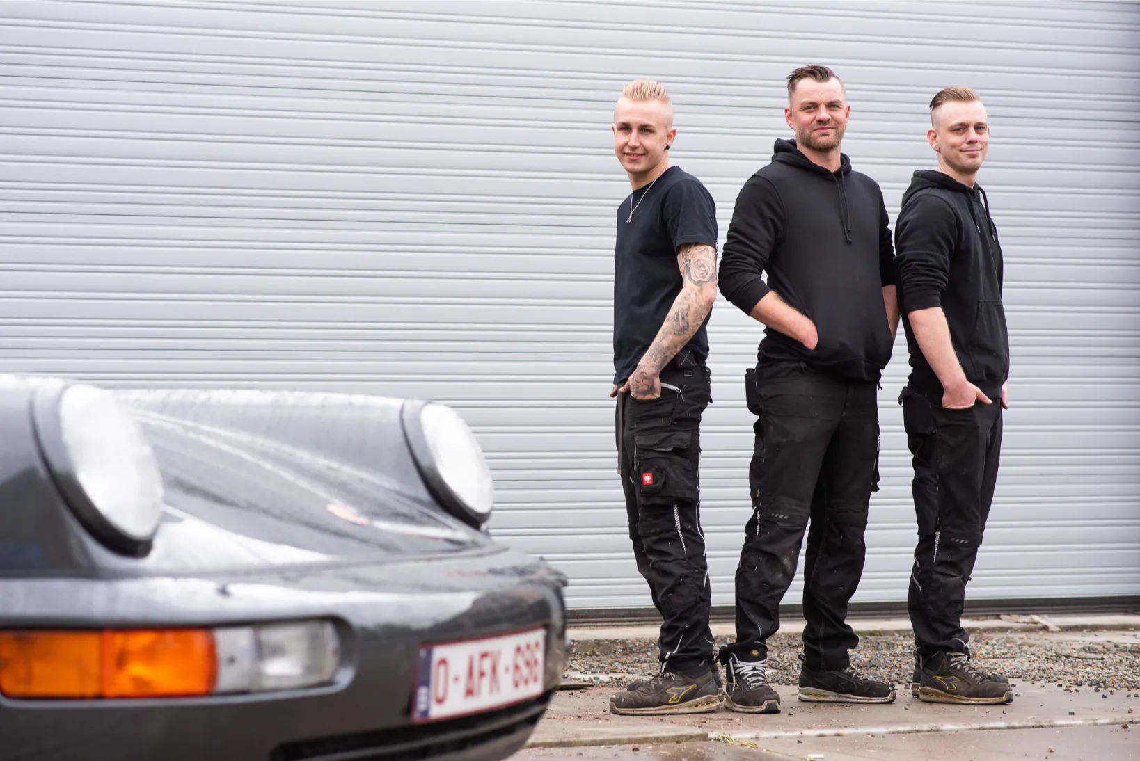 Drietal mannen uit een autobedrijf poserend voor hun loods, met sportwagen op de voorgrond