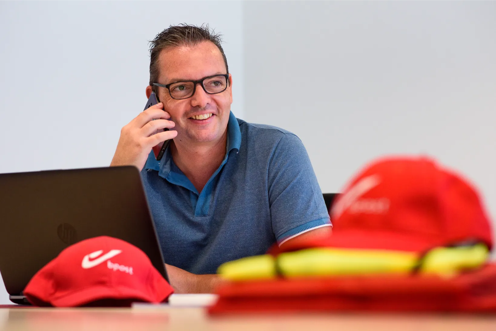Authentiek moment op kantoor: man belt met glimlach achter zijn laptop, met branding zichtbaar op tafel.