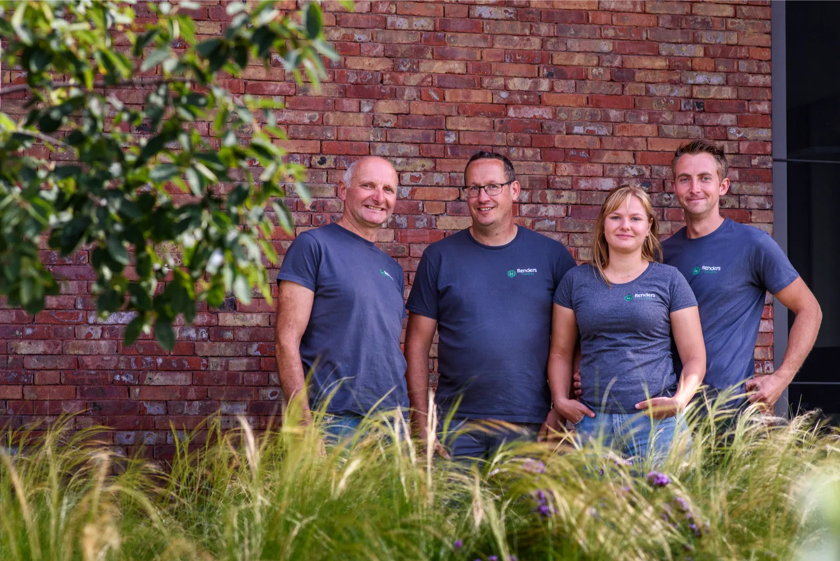 Teamfoto van vier werknemers buiten tegen een bakstenen muur, casual en benaderbaar in beeld gebracht.