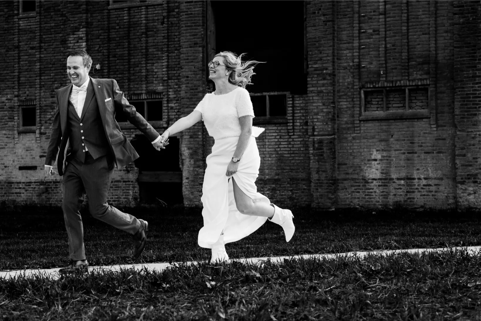 Bruidskoppel dat lachend hand in hand wegloopt bij een industriële locatie — puur en dynamisch.