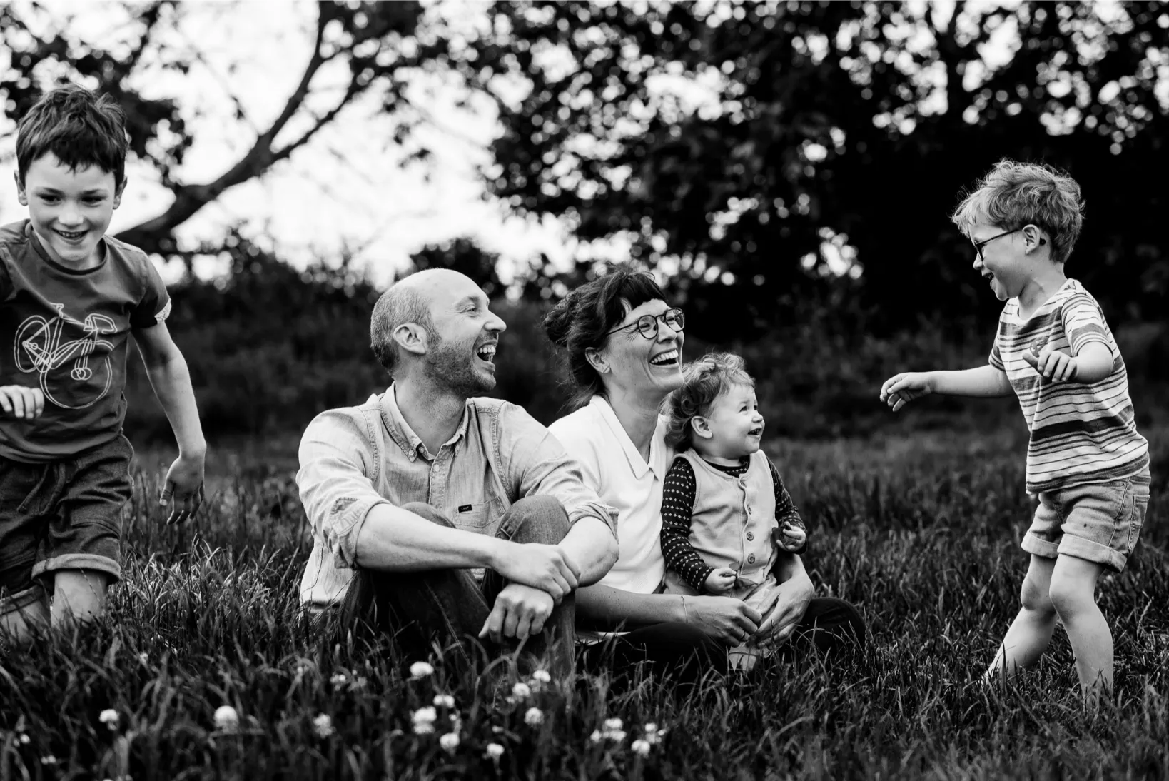Ongeposeerde familiefoto in zwart-wit – ouders en kinderen lachen en spelen in het gras tijdens reportage.